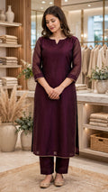 A woman modeling a deep purple three-piece kurta set with matching pants in a boutique setting. The set features a solid purple cotton kurta with sheer purple sleeves and a split neckline, decorated with gold zari borders.