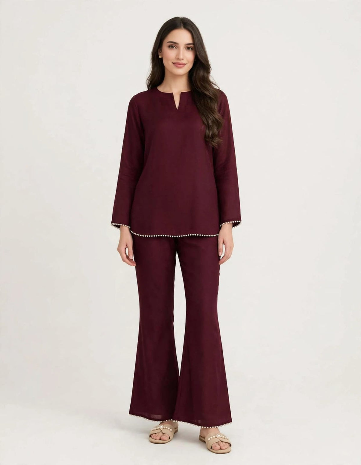A woman modeling a matching deep burgundy two-piece set consisting of a long-sleeve tunic top with a split neckline and matching flared pants, styled with neutral sandals against a plain light background.